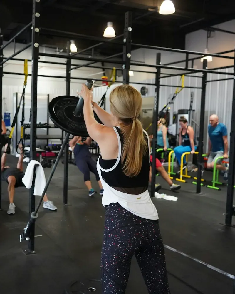 Woman pressing a weighted barbell overhead during a group bootcamp style workout while other members train on rigs and benches behind her