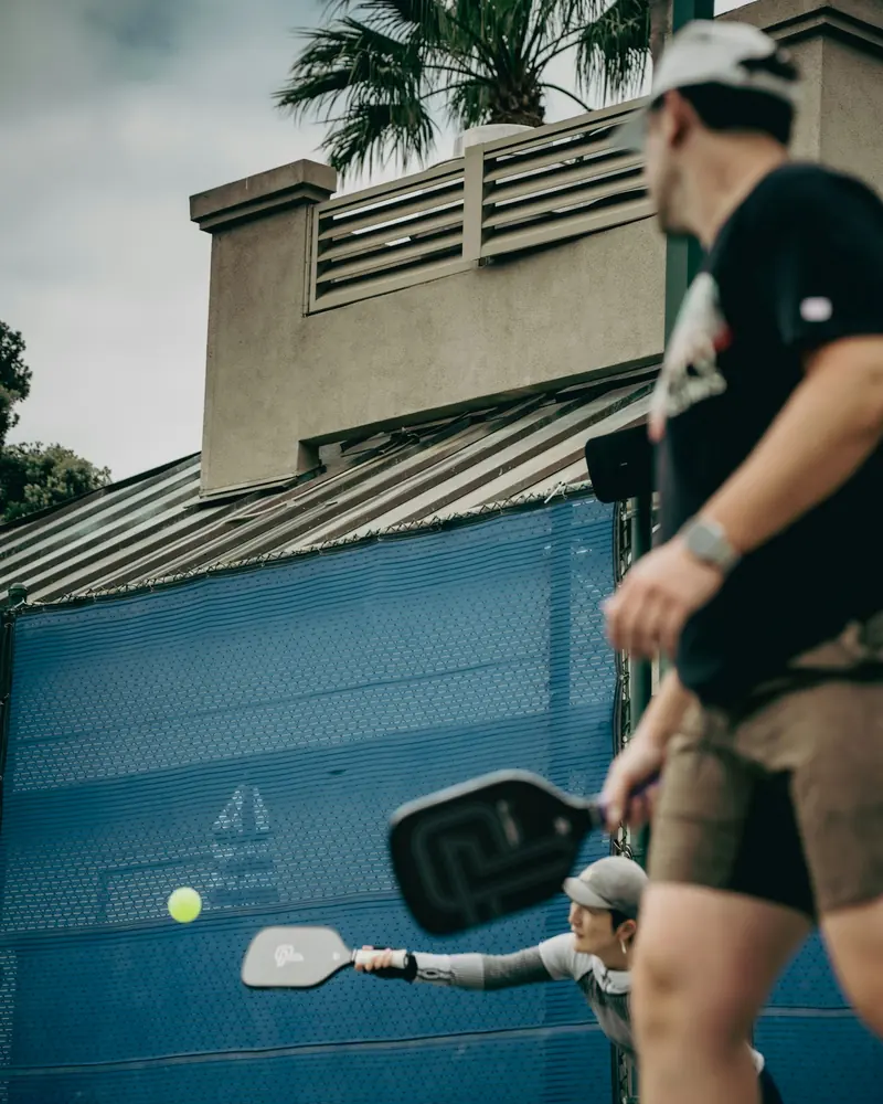 Two people playing doubles pickleball on an outdoor court, one reaching for a low ball near the net, with a palm tree in the background